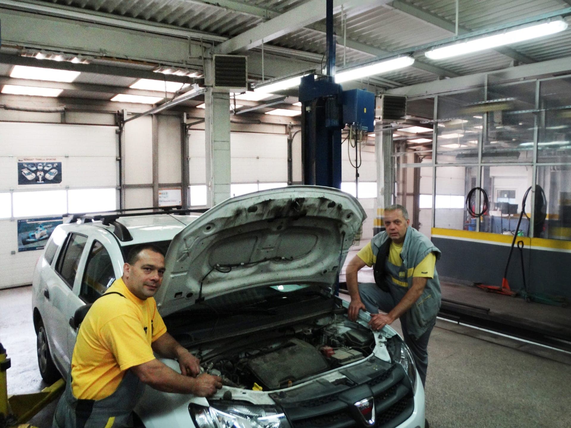 Двама механици в жълто-сиви работни дрехи до бяла Dacia с отворен преден капак в цеха